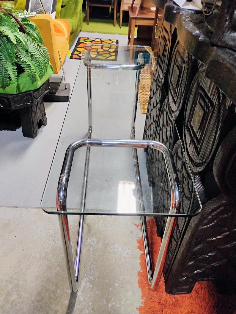 Peut inclure: Table d'appoint vintage en chrome et verre, avec deux &eacute;tag&egrave;res rectangulaires en verre. La table a un design &eacute;pur&eacute; et minimaliste avec des pieds et des supports chrom&eacute;s incurv&eacute;s. Les &eacute;tag&egrave;res en verre sont transparentes, offrant une esth&eacute;tique moderne.