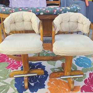 May include: Two vintage swivel bar stools with cream-colored padded seats and backs. The wooden frames have a warm, natural finish. The stools sit on a colorful floral rug.