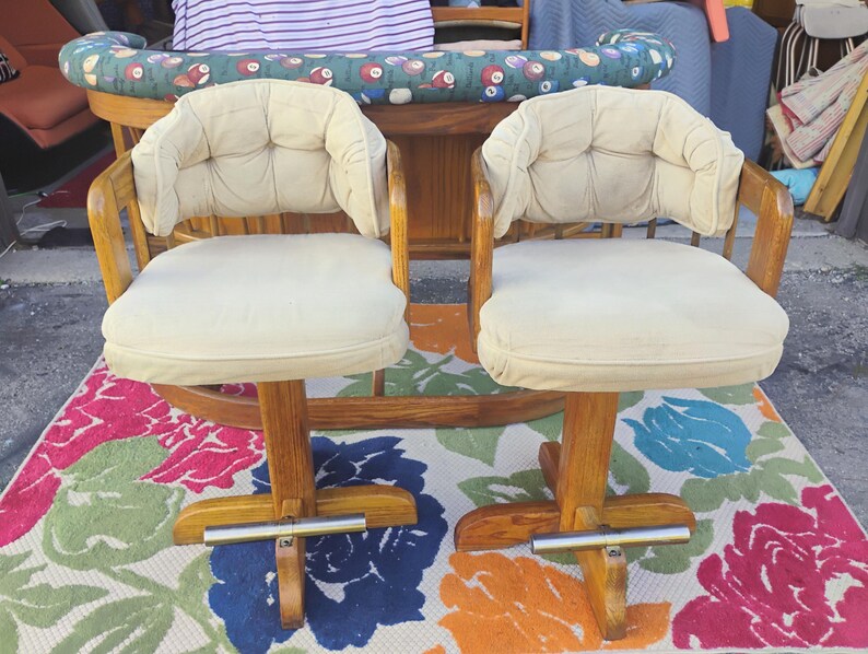 May include: Two vintage swivel bar stools with cream-colored padded seats and backs. The wooden frames have a warm, natural finish. The stools sit on a colorful floral rug.
