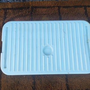 May include: A rectangular white plastic storage bin lid featuring a series of vertical parallel ridges across the surface, a centered circular indentation, and two elongated rectangular slots positioned on the left and right sides.