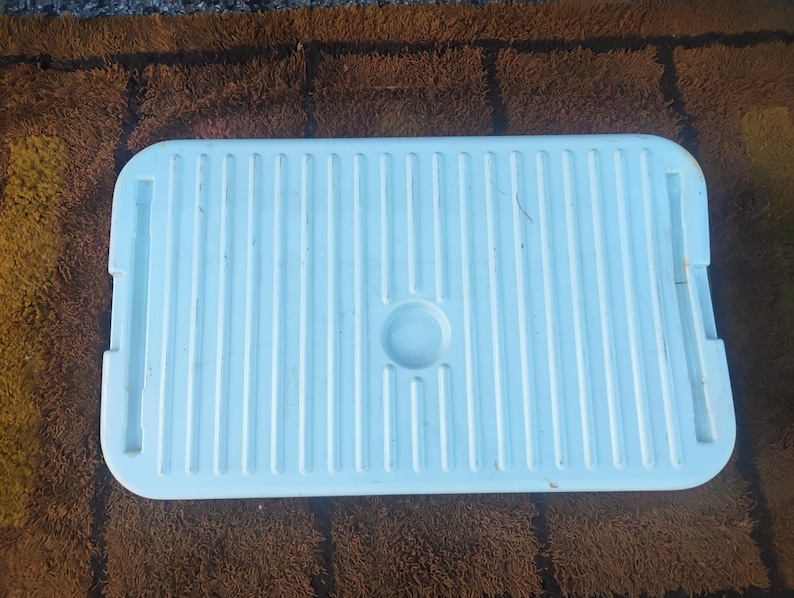 May include: A rectangular white plastic storage bin lid featuring a series of vertical parallel ridges across the surface, a centered circular indentation, and two elongated rectangular slots positioned on the left and right sides.