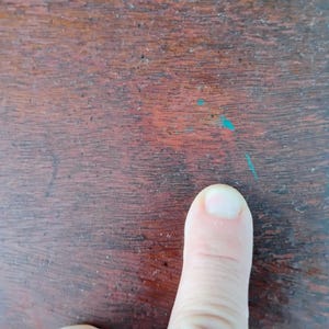 Puede incluir: Primer plano de una superficie de madera marr&oacute;n oscuro con vetas visibles y peque&ntilde;as manchas de pintura turquesa. Un dedo humano apunta hacia la superficie.
