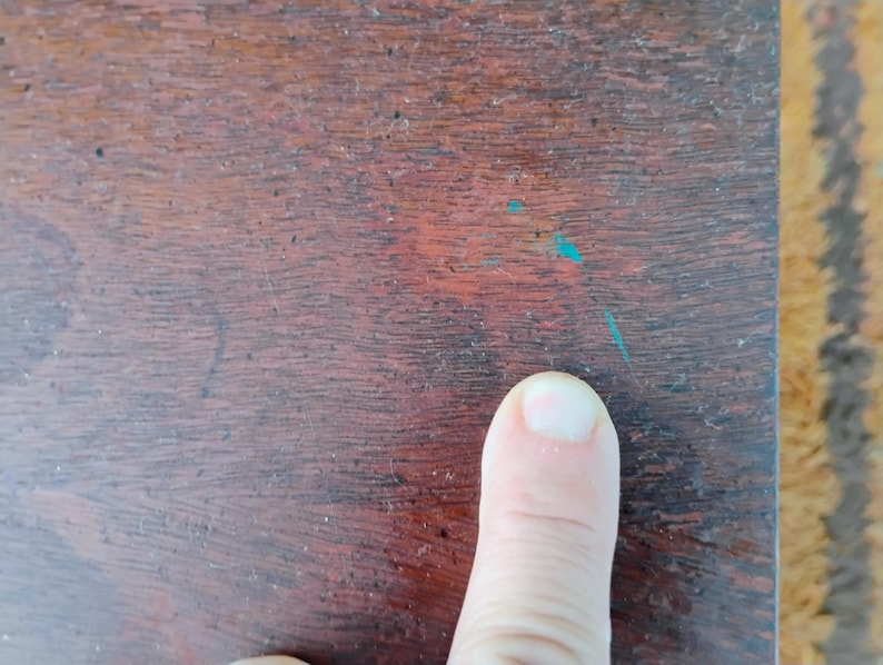 Puede incluir: Primer plano de una superficie de madera marr&oacute;n oscuro con vetas visibles y peque&ntilde;as manchas de pintura turquesa. Un dedo humano apunta hacia la superficie.