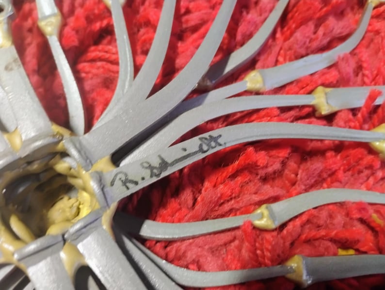 May include: Close-up of a gray and yellow sculpture with curved, blade-like elements. The sculpture is set against a vibrant red, textured background. The artist's signature, "R. Schmidt", is visible on one of the gray elements.
