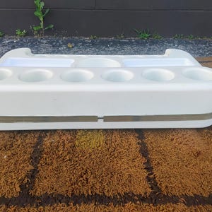May include: A white plastic organizer tray featuring six circular cup holders along the front edge, two rectangular compartments, and one central circular indentation on the top surface, accented by a horizontal translucent band around the middle.
