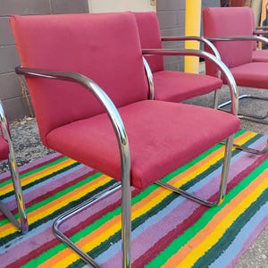 May include: A set of armchairs featuring deep red fabric upholstery on the seat and backrest, supported by a continuous, curved chrome metal frame that forms the armrests and the base. The chairs have a structured, boxy design with clean lines.