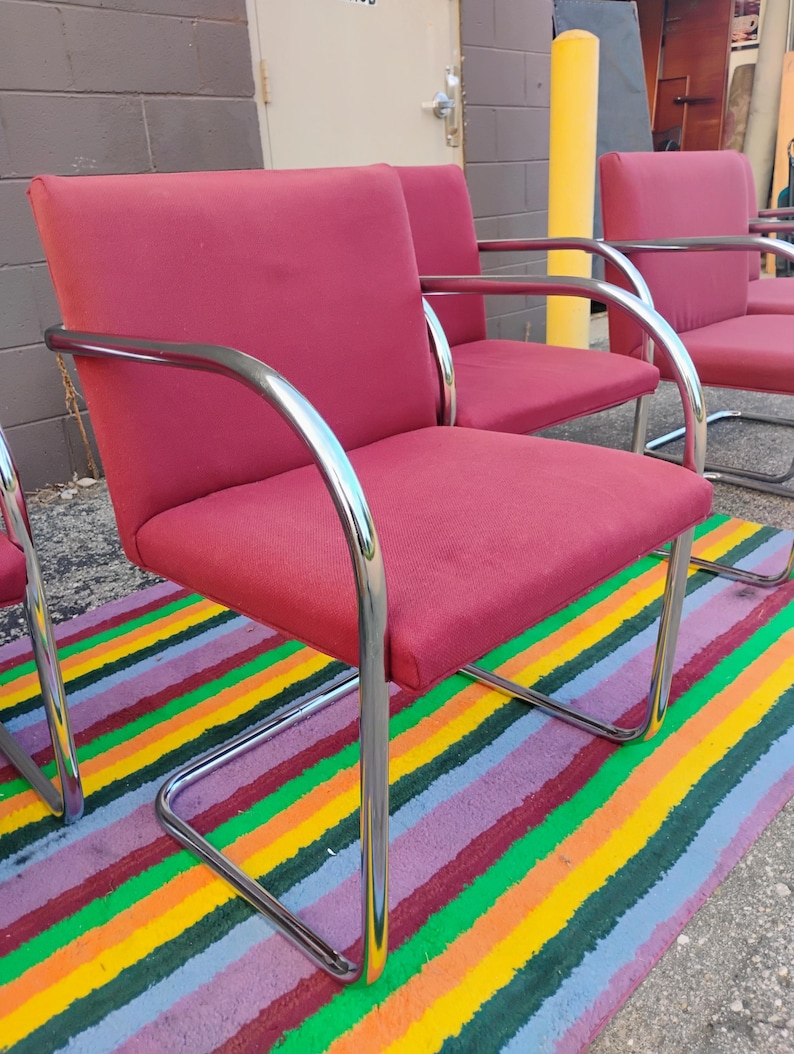 May include: A set of armchairs featuring deep red fabric upholstery on the seat and backrest, supported by a continuous, curved chrome metal frame that forms the armrests and the base. The chairs have a structured, boxy design with clean lines.