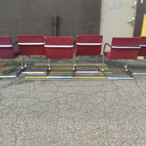 May include: A set of six red chairs with rectangular backrests and seats, each featuring a continuous chrome metal cantilever frame. The chairs are arranged in a row, showcasing a uniform design with horizontal chrome bars across the backrests.