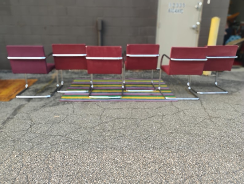May include: A set of six red chairs with rectangular backrests and seats, each featuring a continuous chrome metal cantilever frame. The chairs are arranged in a row, showcasing a uniform design with horizontal chrome bars across the backrests.