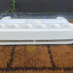 May include: A white plastic organizer tray featuring six circular cup holders along the front edge, two rectangular compartments, and one central circular indentation on the top surface, accented by a horizontal translucent band around the middle.