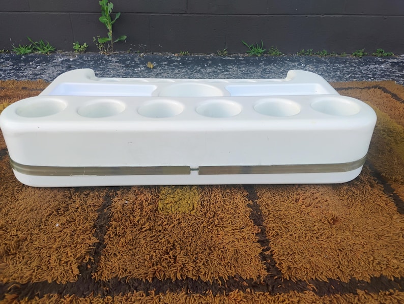 May include: A white plastic organizer tray featuring six circular cup holders along the front edge, two rectangular compartments, and one central circular indentation on the top surface, accented by a horizontal translucent band around the middle.