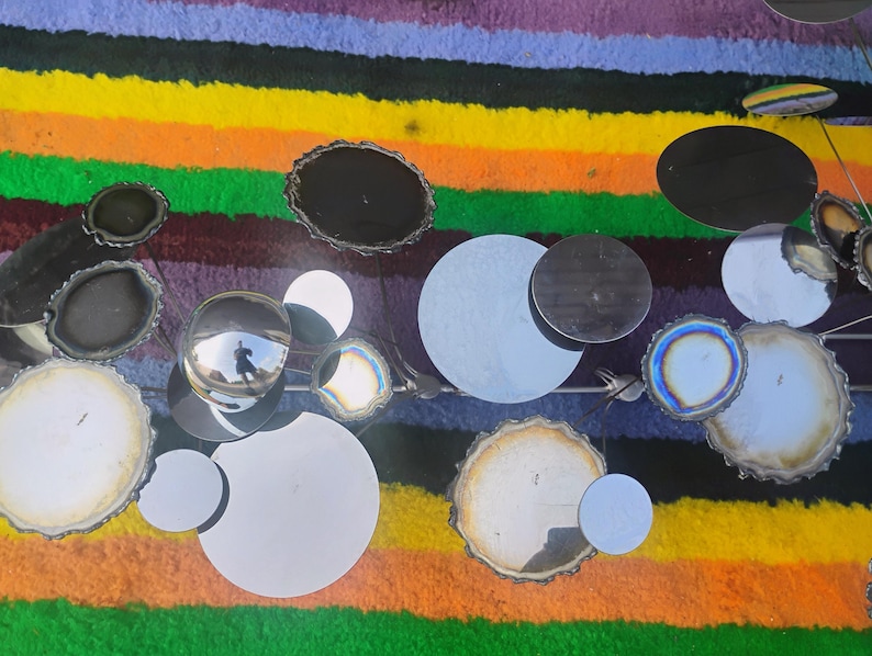 May include: A wall sculpture featuring a collection of circular mirrors and polished agate slices in shades of black, white, and iridescent blue. The pieces are arranged on a metal frame, creating a reflective and textured composition of varied sizes.