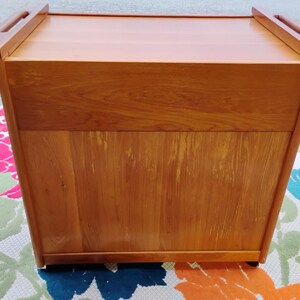 May include: A wooden serving cart with a lift-up top and wheels. The cart is made of light brown wood and has a simple design.