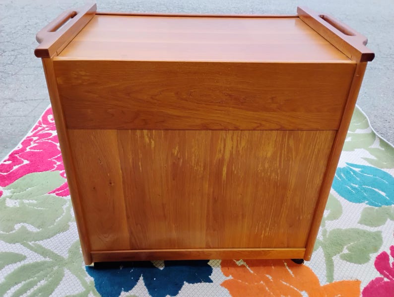May include: A wooden serving cart with a lift-up top and wheels. The cart is made of light brown wood and has a simple design.