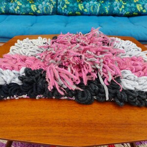 May include: A handmade rug with a textured design. The rug features a combination of pink, gray, and black yarn. The rug is placed on a wooden table, with a blue couch in the background.