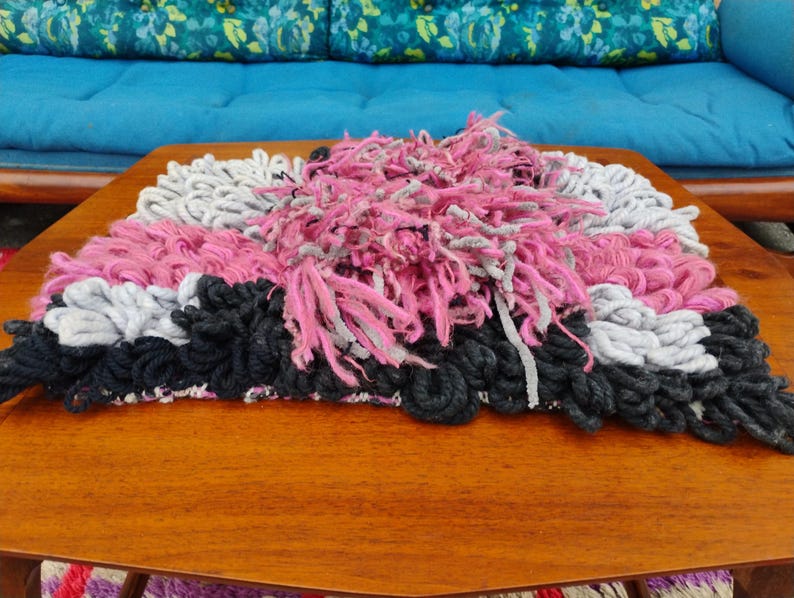 May include: A handmade rug with a textured design. The rug features a combination of pink, gray, and black yarn. The rug is placed on a wooden table, with a blue couch in the background.