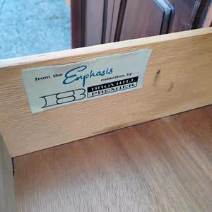 May include: The interior side of a wooden drawer featuring a rectangular label that reads from the Emphasis collection by Broyhill Premier with a stylized logo. The light-colored wood grain is visible on the side panel and the bottom surface.