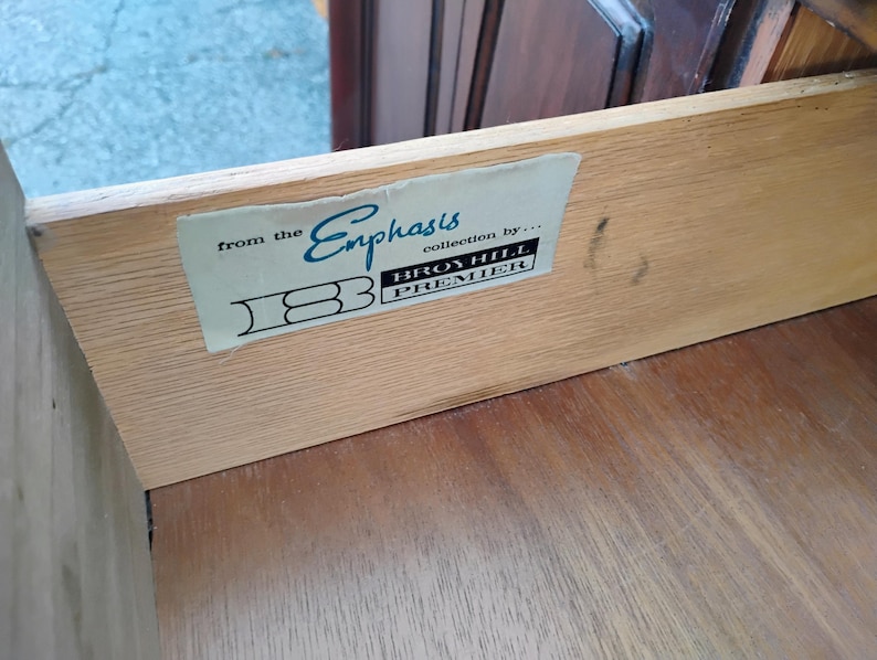 May include: The interior side of a wooden drawer featuring a rectangular label that reads from the Emphasis collection by Broyhill Premier with a stylized logo. The light-colored wood grain is visible on the side panel and the bottom surface.