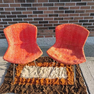 May include: Two mid-century modern chairs with vibrant orange fabric seats and slender metal legs. The chairs are set on a multi-colored shag rug, with a brick wall in the background. The chairs have a unique, curved back design.