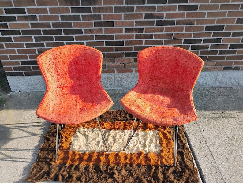 May include: Two mid-century modern chairs with vibrant orange fabric seats and slender metal legs. The chairs are set on a multi-colored shag rug, with a brick wall in the background. The chairs have a unique, curved back design.