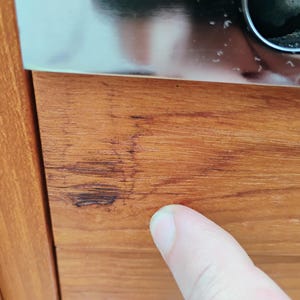 May include: A close-up view of a smooth, light brown wooden surface featuring a distinct, dark brown linear scratch or blemish running horizontally across the grain. The wood displays a natural, warm-toned finish with visible vertical grain patterns.