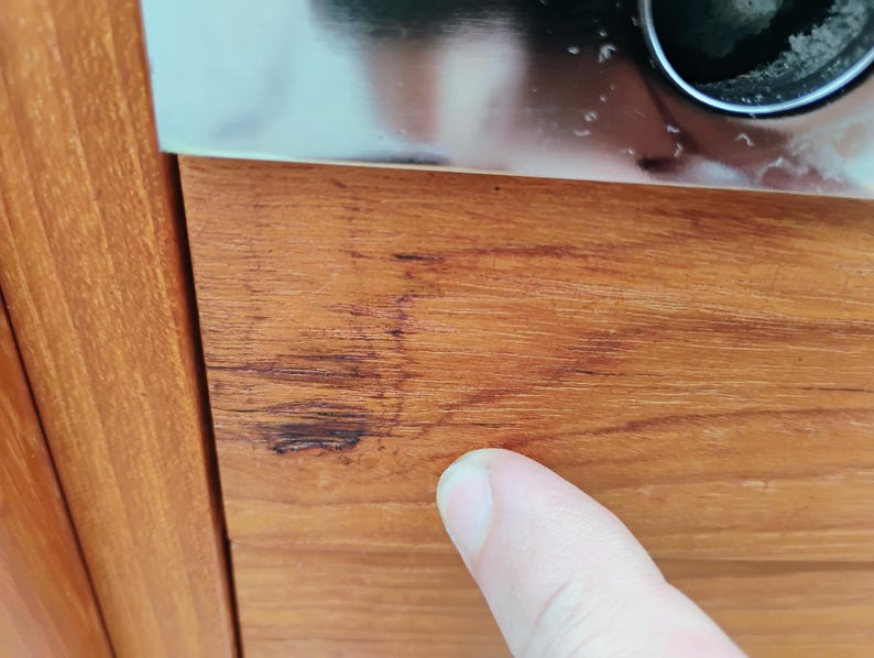 May include: A close-up view of a smooth, light brown wooden surface featuring a distinct, dark brown linear scratch or blemish running horizontally across the grain. The wood displays a natural, warm-toned finish with visible vertical grain patterns.