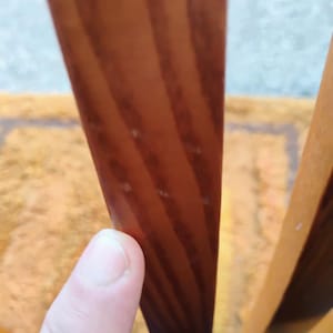 May include: A vertical wooden slat with a smooth, polished finish and visible brown wood grain patterns. The slat is positioned upright, showcasing the natural texture and warm, medium-toned color of the wood material in a clear, focused view.