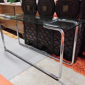 Peut inclure: Une console rectangulaire dot&eacute;e d'un plateau en verre transparent soutenu par une structure tubulaire continue en m&eacute;tal chrom&eacute;. Le cadre forme une base &eacute;l&eacute;gante aux coins arrondis, offrant une structure stable pour la surface en verre.