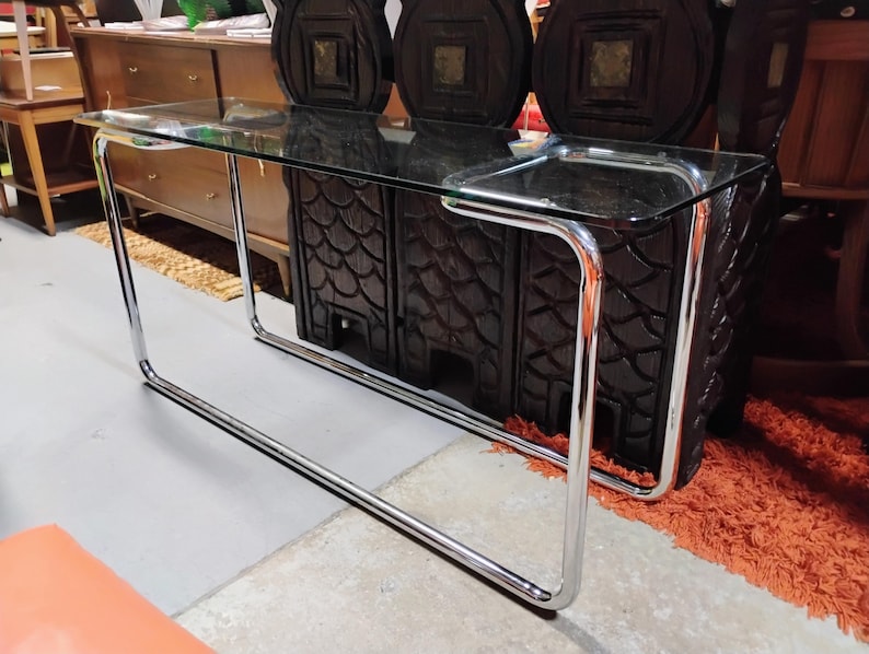 Peut inclure: Une console rectangulaire dot&eacute;e d'un plateau en verre transparent soutenu par une structure tubulaire continue en m&eacute;tal chrom&eacute;. Le cadre forme une base &eacute;l&eacute;gante aux coins arrondis, offrant une structure stable pour la surface en verre.