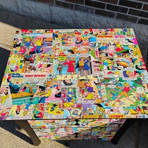 May include: A black, three-drawer chest with a comic book collage design. The top and drawer fronts are covered in colorful comic book panels featuring various characters and speech bubbles. The chest has black legs and a rectangular shape.