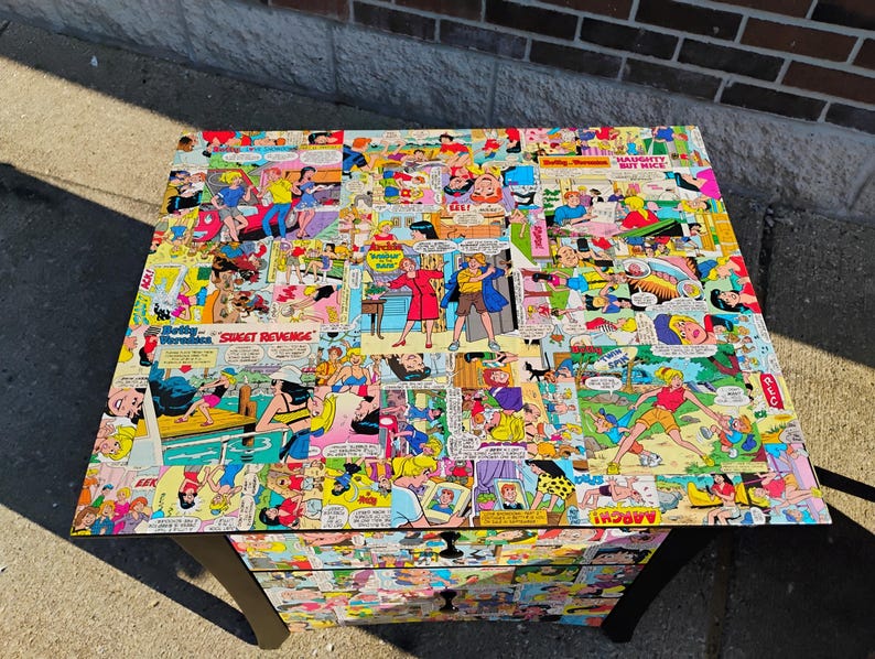 May include: A black, three-drawer chest with a comic book collage design. The top and drawer fronts are covered in colorful comic book panels featuring various characters and speech bubbles. The chest has black legs and a rectangular shape.