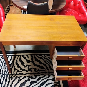 May include: A wooden desk with a light brown finish. The desk has a rectangular top and three drawers on the right side. The drawers are open, revealing a dark interior. The desk is supported by four legs. The desk is on a black and white zebra print rug.