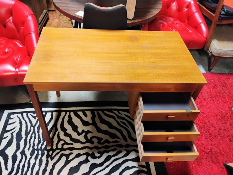 May include: A wooden desk with a light brown finish. The desk has a rectangular top and three drawers on the right side. The drawers are open, revealing a dark interior. The desk is supported by four legs. The desk is on a black and white zebra print rug.
