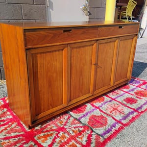 Op de afbeelding: Een rechthoekig houten dressoir met twee bovenste lades met verzonken grepen en vier onderste kastdeuren. Het meubel is gemaakt van warmgekleurd hout met een zichtbare houtnerf en staat op een platte basis met een strak, lineair ontwerp.