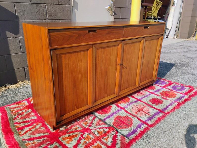 Op de afbeelding: Een rechthoekig houten dressoir met twee bovenste lades met verzonken grepen en vier onderste kastdeuren. Het meubel is gemaakt van warmgekleurd hout met een zichtbare houtnerf en staat op een platte basis met een strak, lineair ontwerp.