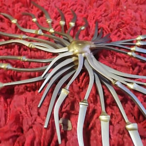 May include: A decorative metal sunburst sculpture with a central gold-colored sphere and radiating silver-colored arms. The arms have gold-colored accents and are arranged against a red textured background. The sculpture has a unique, artistic design.