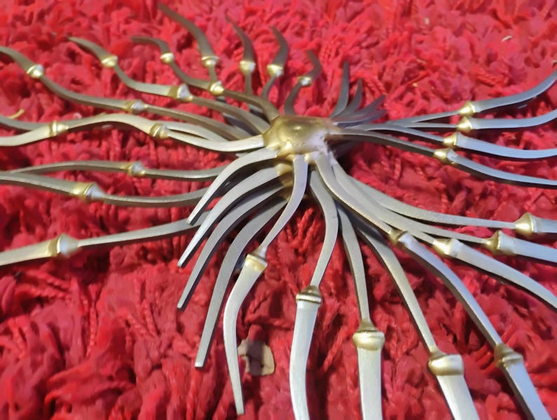 May include: A decorative metal sunburst sculpture with a central gold-colored sphere and radiating silver-colored arms. The arms have gold-colored accents and are arranged against a red textured background. The sculpture has a unique, artistic design.
