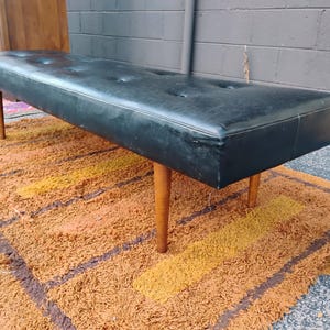 May include: A long, rectangular bench featuring a black tufted seat cushion. The bench is supported by four tapered, light-colored wooden legs. The seat surface has a smooth texture with visible button detailing across the top, providing a flat, elongated profile for seating.