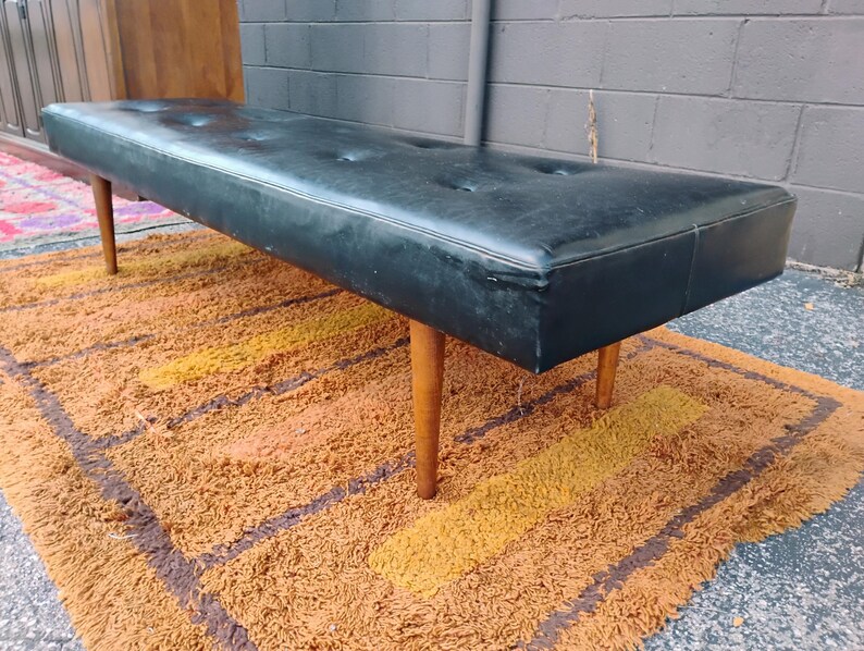 May include: A long, rectangular bench featuring a black tufted seat cushion. The bench is supported by four tapered, light-colored wooden legs. The seat surface has a smooth texture with visible button detailing across the top, providing a flat, elongated profile for seating.