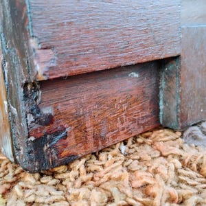 Puede incluir: Primer plano de una esquina de una caja de madera, mostrando la carpinter&iacute;a y la madera envejecida, de color marr&oacute;n oscuro. La caja descansa sobre una superficie texturizada, de color marr&oacute;n claro y naranja, con desgaste visible.