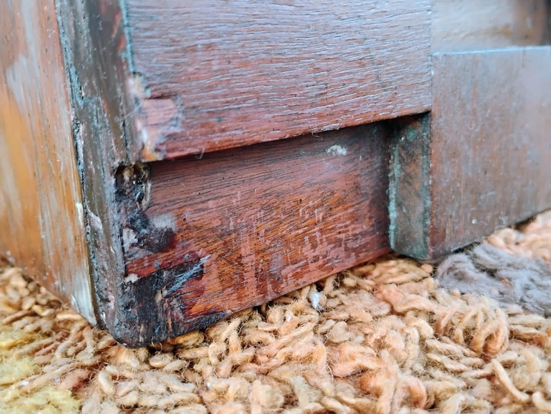 Puede incluir: Primer plano de una esquina de una caja de madera, mostrando la carpinter&iacute;a y la madera envejecida, de color marr&oacute;n oscuro. La caja descansa sobre una superficie texturizada, de color marr&oacute;n claro y naranja, con desgaste visible.