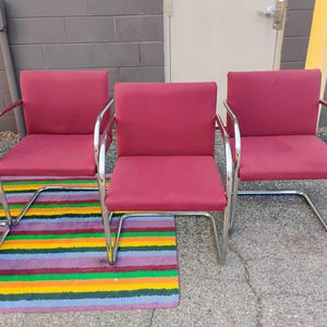 May include: A set of three armchairs featuring deep red upholstered seats and backrests. Each chair is supported by a continuous, polished chrome tubular metal frame that forms a cantilevered base, providing a sleek and structural appearance.