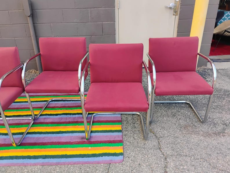 May include: A set of three armchairs featuring deep red upholstered seats and backrests. Each chair is supported by a continuous, polished chrome tubular metal frame that forms a cantilevered base, providing a sleek and structural appearance.