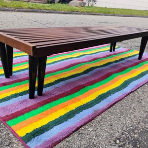 K&ouml;nnte beinhalten: Eine Bank im Mid-Century-Stil mit einer Lattenoberfl&auml;che aus Holz und schwarzen, sich verj&uuml;ngenden Beinen. Die Bank steht auf einem bunten Teppich mit horizontalen Streifen in verschiedenen Farben, darunter Rot, Orange, Gelb, Gr&uuml;n und Lila.