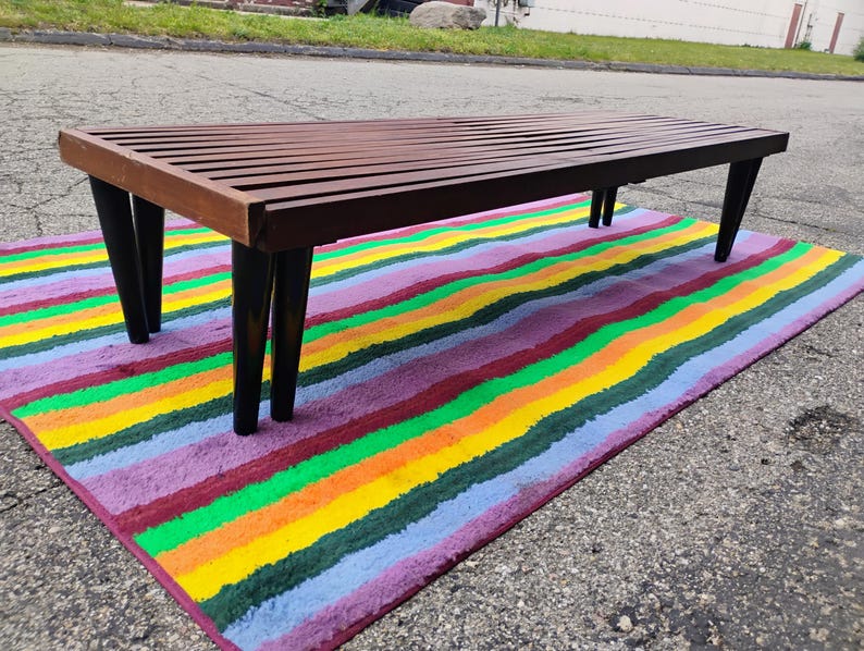 K&ouml;nnte beinhalten: Eine Bank im Mid-Century-Stil mit einer Lattenoberfl&auml;che aus Holz und schwarzen, sich verj&uuml;ngenden Beinen. Die Bank steht auf einem bunten Teppich mit horizontalen Streifen in verschiedenen Farben, darunter Rot, Orange, Gelb, Gr&uuml;n und Lila.