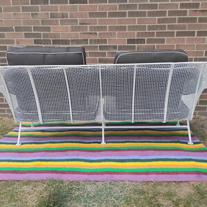 May include: A white metal wire sofa featuring a grid pattern frame with two dark gray cushions. The sofa has a high backrest and thin metal legs, providing a structured seating design. The cushions are rectangular with white piping along the edges.