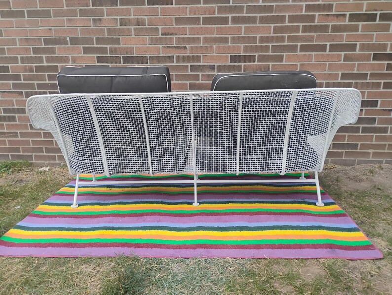 May include: A white metal wire sofa featuring a grid pattern frame with two dark gray cushions. The sofa has a high backrest and thin metal legs, providing a structured seating design. The cushions are rectangular with white piping along the edges.