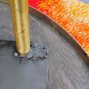 Op de afbeelding: Close-up van een zwarte ronde tafelbasis met een messing paal en een zwarte metalen beugel. De tafelbasis is bevestigd aan een zwarte metalen rand.