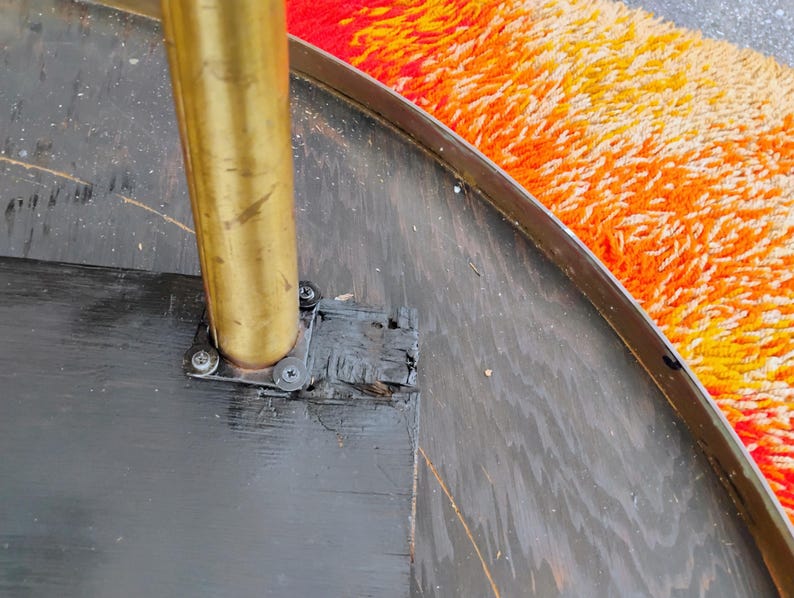 Op de afbeelding: Close-up van een zwarte ronde tafelbasis met een messing paal en een zwarte metalen beugel. De tafelbasis is bevestigd aan een zwarte metalen rand.