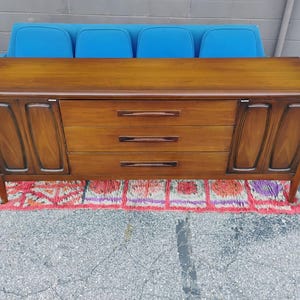 May include: A long wooden sideboard featuring a smooth rectangular top, three central horizontal drawers with dark wood handles, and two side cabinets with recessed panel doors. The piece stands on four tapered legs and has a warm brown finish.